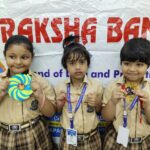 Three young students in beige uniforms pose with handmade crafts in front of a raksha bandhan banner